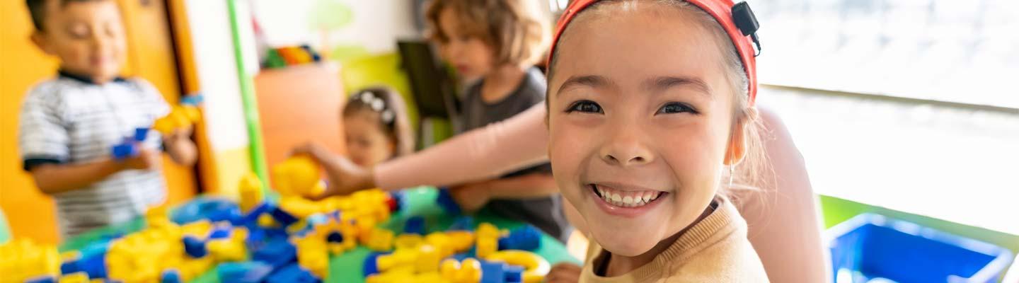 Young girl smiles at the camera in front of a table with blocks, a staff member and other children