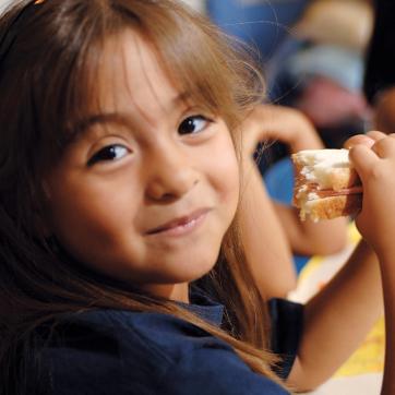 Preschooler smiling at the camera as she eats her sandwich