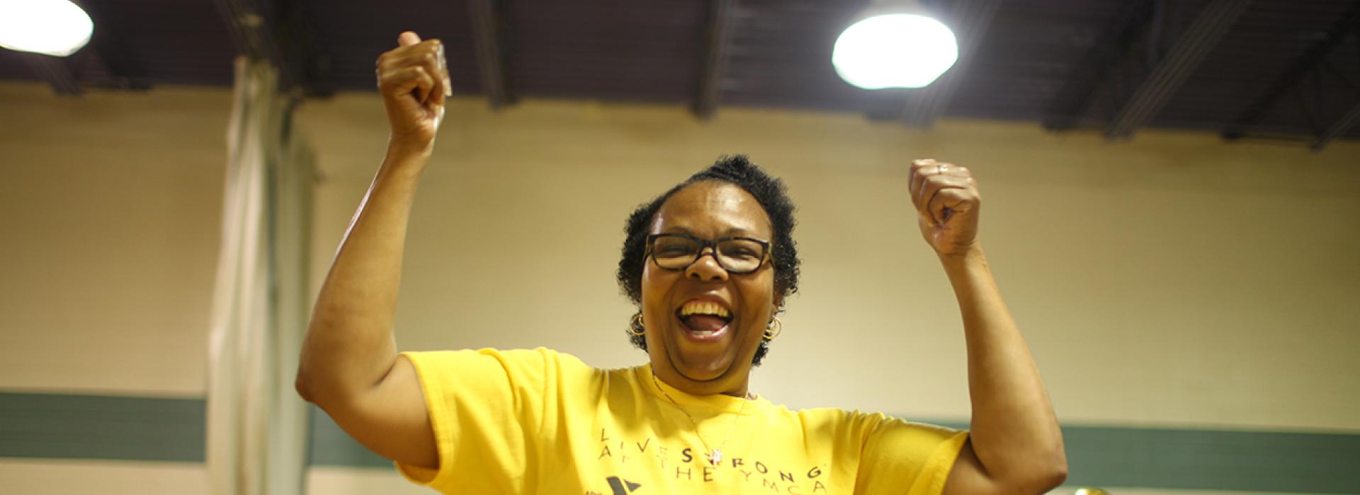 A Livestrong at the YMCA participant and cancer survivor smiles at the camera with arms in the air, cheering